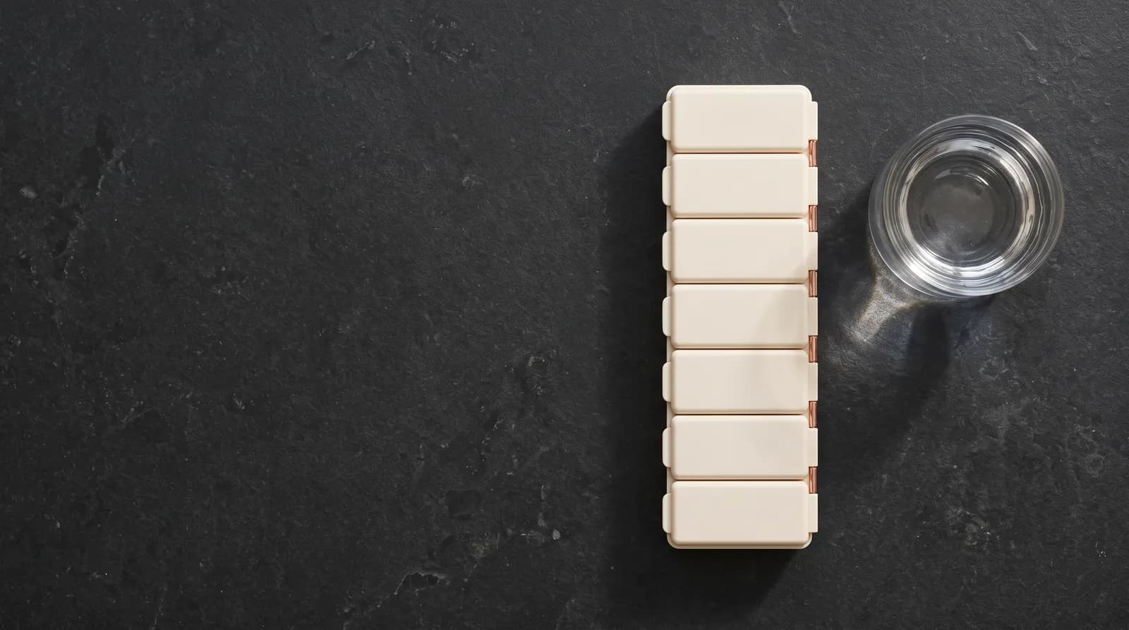 A cream weekly organizer with closed lids beside a clear glass of water on dark stone, seen from above.